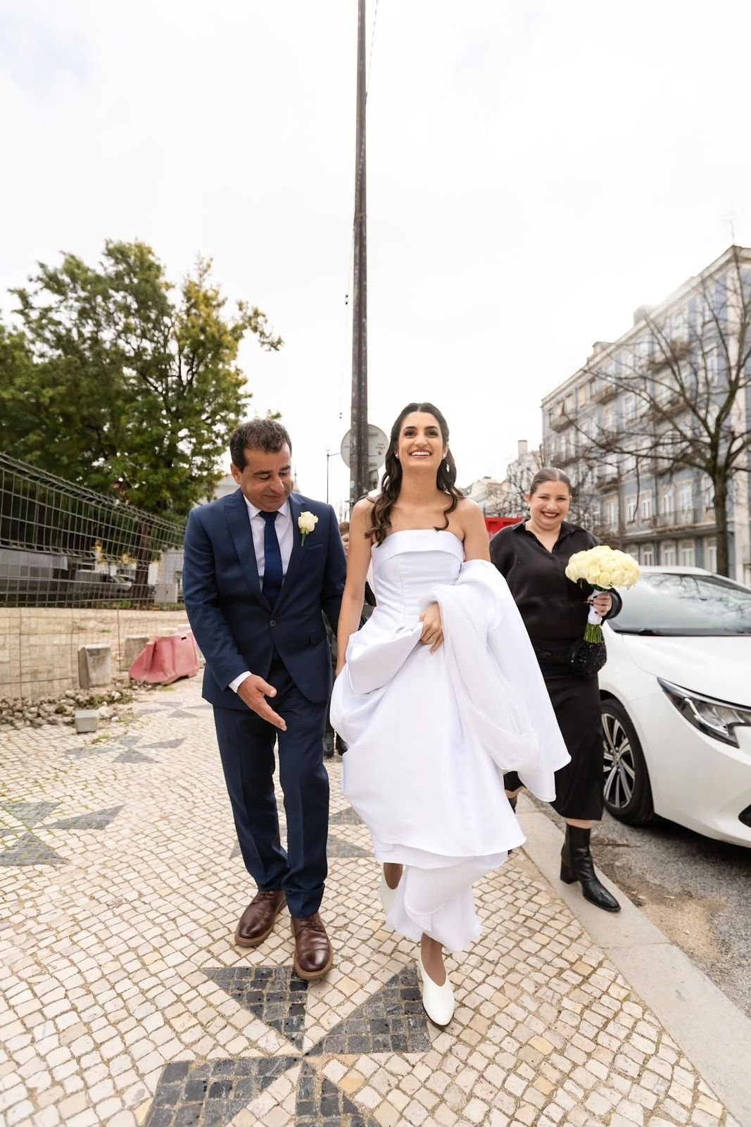 Noiva a caminhar na calçada portuguesa com o pai, seguida pela wedding planner com o ramo de rosas brancas