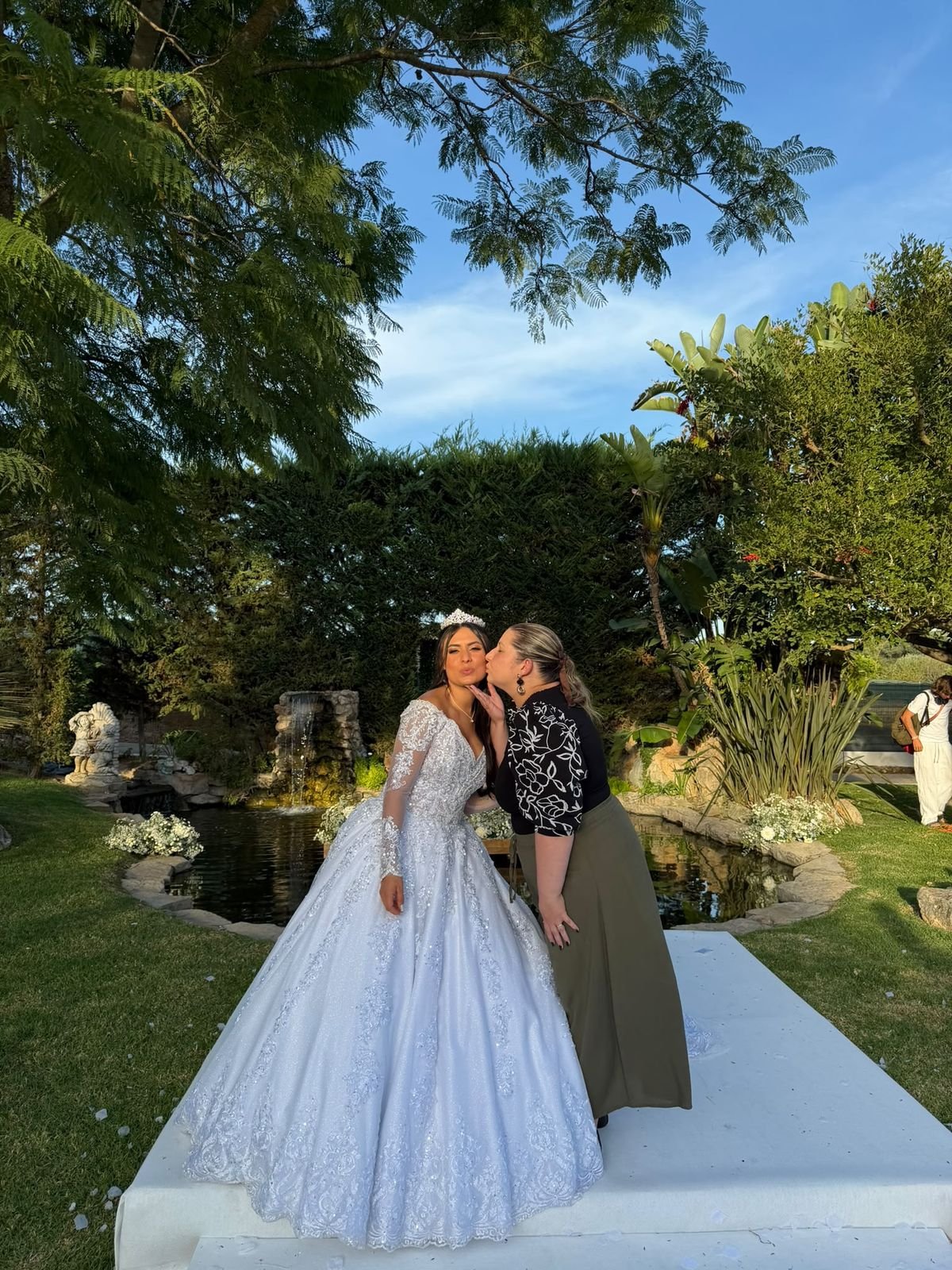 Noiva de vestido de princesa a receber um beijo na bochecha da wedding planner num jardim com lago