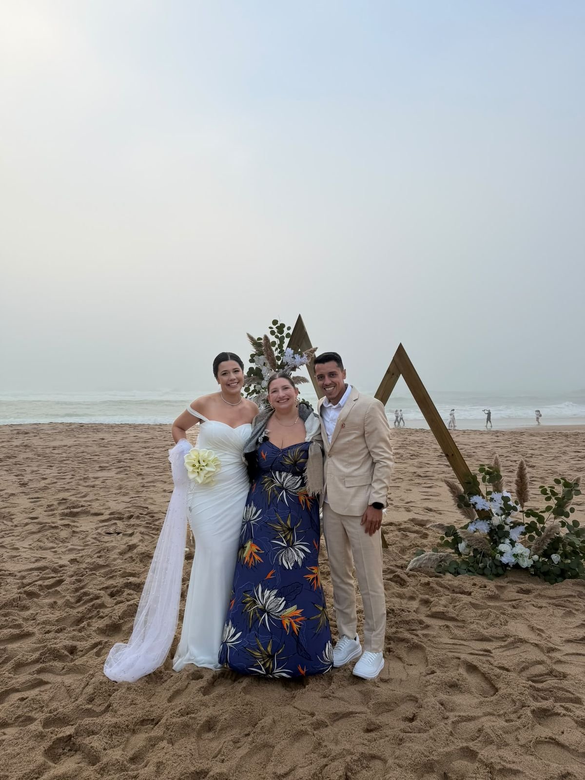 Noivos com a wedding planner numa cerimónia na praia, junto a um arco triangular de madeira com flores